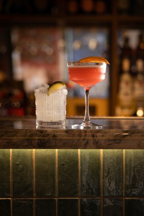 A tall, pink cocktail with a slice of orange next to a small white cocktail with a slice of lime, sat on the bar in Banksia Leeds