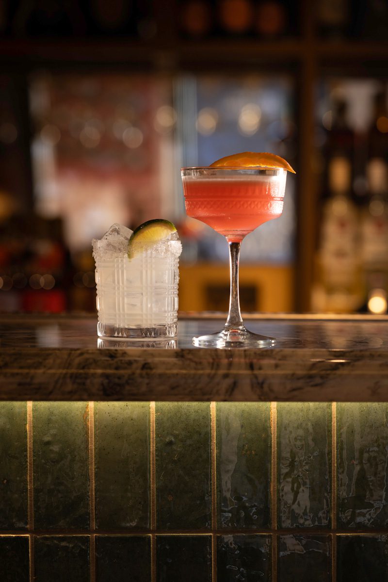 A tall, pink cocktail with a slice of orange next to a small white cocktail with a slice of lime, sat on the bar in Banksia Leeds